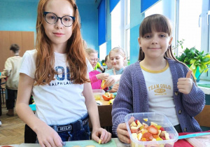 Dwie uczennice prezentują sałatkę owocową w miseczce.
