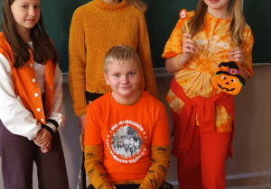 Czwórka uczniów w pomarańczowych ubraniach. Jeden chłopiec siedzi.