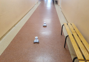 Dwa roboty na szkolnym korytarzu. Po prawej stronie stoi ławka.