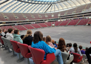 Dzieci siedzą tyłem na trybunach stadionu.
